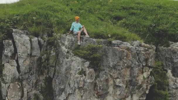 Giovane donna hipster è seduto sul bordo sull'erba di alto muro di roccia. Vista aerea. Drone sta volando lento intorno dal modello — Video Stock