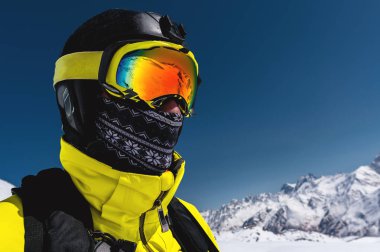 Bir kayakçı maskesi ve kask karla kaplı dağlar ve mavi gökyüzü bir arka plan karşı kapalı bir yüz ile yakın çekim portre