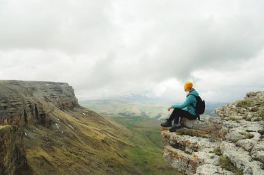 Genç kadın ikindilerde kaya kenarında oturan ve bulutlar ile gökyüzüne bakarak bir sırt çantası ile