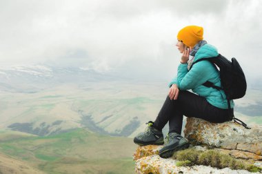 Genç kadın ikindilerde kaya kenarında oturan ve bulutlar ile gökyüzüne bakarak bir sırt çantası ile
