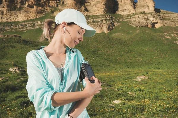 Спортивна жінка в шапці і нужники перемикає пісні на свій мобільний телефон під час вправ на відкритому повітрі на тлі полів і каменів — стокове фото