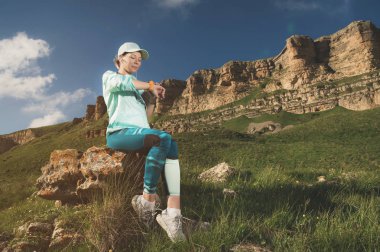 Bir kap ve kayalar bir arka plan açık havada bir kayaya oturan akıllı saatini kontrol kulaklıklar bir gülümseyen genç fitness kız portresi