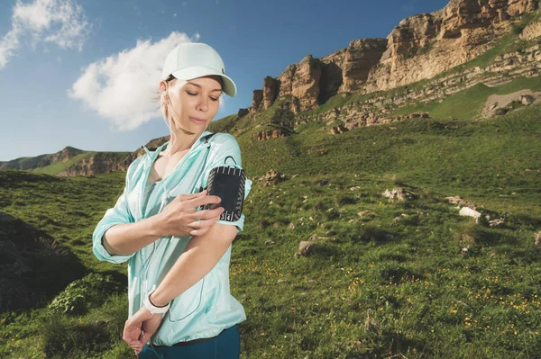 Uma mulher de esportes em um boné e nuzhniki muda as músicas para seu telefone celular durante um exercício ao ar livre contra o fundo de campos e rochas — Fotografia de Stock