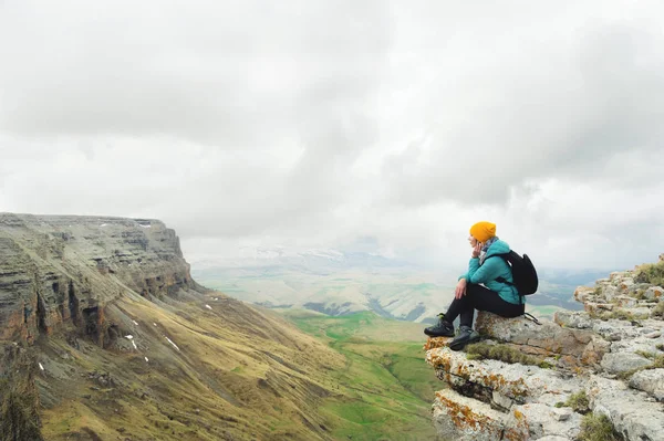 Mujer joven con una mochila pensativamente sentada en el borde de una roca y mirando al cielo con nubes — Foto de Stock