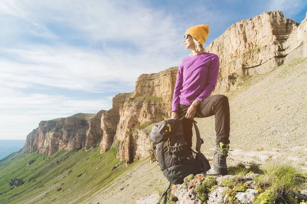 Smiling girl traveler in a yellow hat and a pair of sunglasses stands at the foot of epic rocks with a backpack next and looks away — Stock Photo, Image