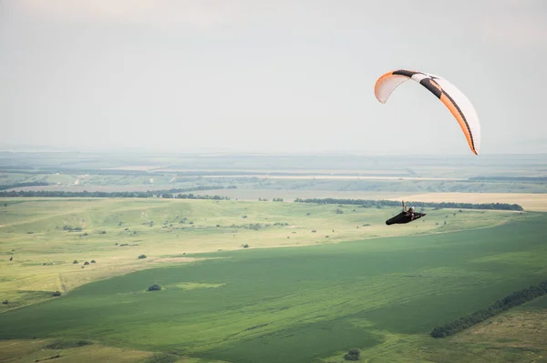 Weiße orange Gleitschirm mit einem Gleitschirm in einem Kokon vor dem Hintergrund der Felder des Himmels und Wolken. Gleitschirmsport — Stockfoto