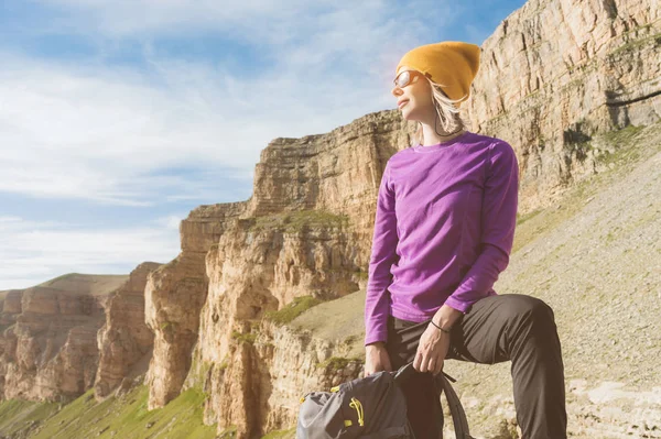 Viajante sorridente menina em um chapéu amarelo e um par de óculos de sol fica ao pé de pedras épicas com uma mochila próxima e olha para longe — Fotografia de Stock