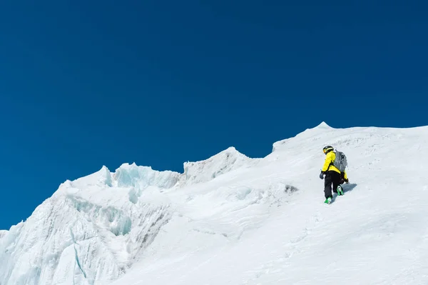 Um esquiador em um capacete e máscara com uma mochila sobe em uma encosta contra o fundo da neve e uma geleira. País de origem Freeride — Fotografia de Stock