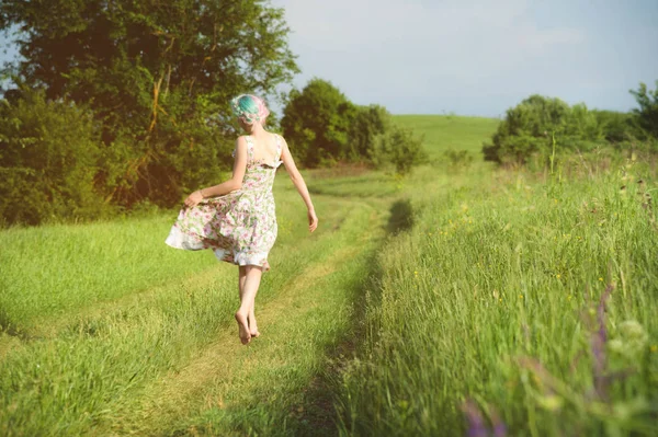 Χαριτωμένο χαρούμενη κοπέλα με ένα ελαφρύ βαμβακερό φόρεμα βόλτες κατά μήκος του δρόμου χώρα με φόντο το καλοκαίρι καταπράσινο τοπίο κατά το ηλιοβασίλεμα. Θέα από το πίσω μέρος — Φωτογραφία Αρχείου