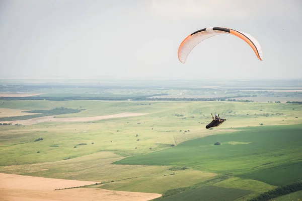 Weiße orange Gleitschirm mit einem Gleitschirm in einem Kokon vor dem Hintergrund der Felder des Himmels und Wolken. Gleitschirmsport — Stockfoto