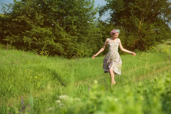 Uma jovem menina alegre bonito em um vestido de algodão luz passeia ao longo da estrada do país contra o pano de fundo da vegetação de verão ao pôr do sol — Fotografia de Stock