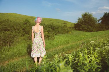 Hafif pamuklu elbise sevimli neşeli genç kız gün batımında yaz yeşil zemin karşı ülke yol boyunca elini kolunu sallaya dönüyor. Arkadan görüntülemek