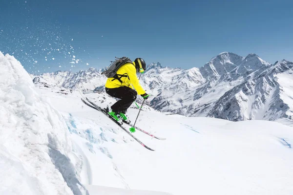 Um esquiador em equipamento desportivo completo salta para o precipício do topo da geleira contra o fundo do céu azul e as montanhas cobertas de neve caucasianas. Região de Elbrus. Rússia — Fotografia de Stock