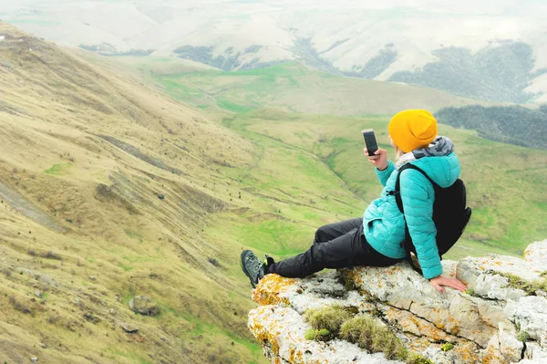 Vista trasera de una mujer sentada en una roca en ropa de invierno y con un sombrero en un teléfono inteligente. Montañas del Cáucaso — Foto de Stock