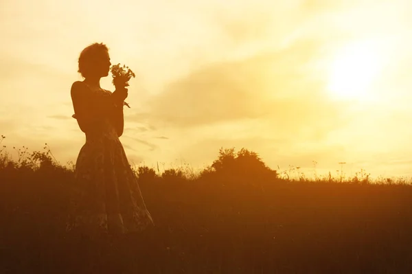 Mujer joven en vestido con ramo de flores en las manos al atardecer en el campo. Imagen de silueta tibia teñida — Foto de Stock