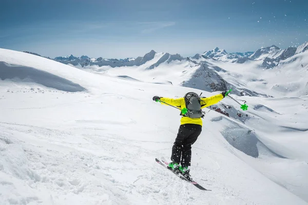 Un esquiador con equipamiento deportivo completo salta al abismo desde la cima del glaciar contra el fondo del cielo azul y las montañas caucásicas cubiertas de nieve. Vista desde atrás. Región de Elbrus — Foto de Stock