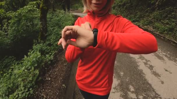 Young attractive woman in headphones changing the settings on a smart watch in front of or psole jogging on the road in a green forest — Stock Video