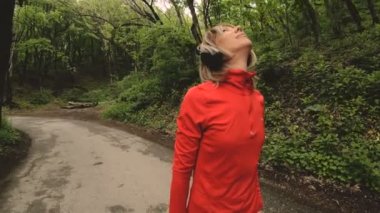 4 k genç sarışın bir kadın Kulaklığımda ormanda çalıştırmadan önce ısınma ve germe gerçekleştirir