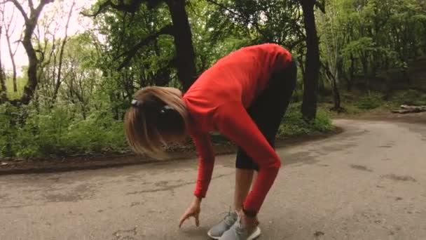 4K joven rubia en auriculares realiza estiramiento y calentamiento antes de correr en el bosque — Vídeo de stock