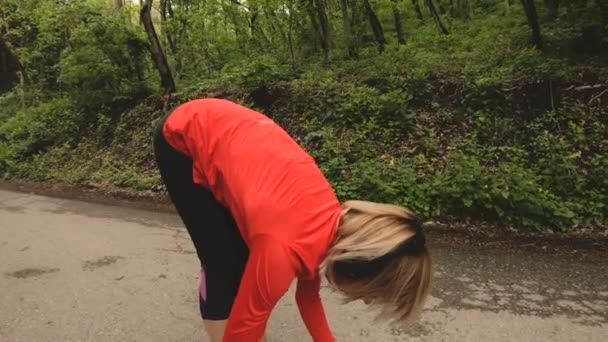 4K jovem loira em fones de ouvido realiza alongamento e aquecimento antes de correr na floresta — Vídeo de Stock