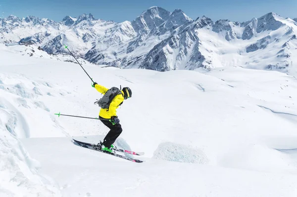 Vor dem Hintergrund des blauen Himmels und der kaukasischen schneebedeckten Berge springt ein Skifahrer in voller Sportausrüstung vom Gipfel des Gletschers in den Abgrund. elbrus region. Russland — Stockfoto