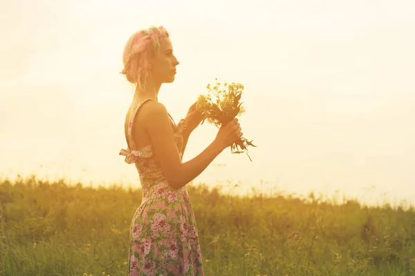 Fiatal nő a ruha, a csokor virág, kezében a naplemente a területen. Színezett meleg kép — Stock Fotó