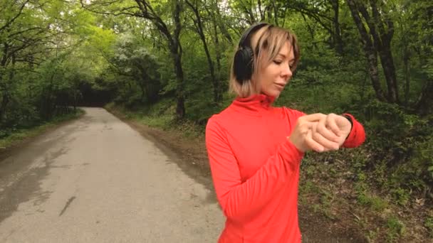 Young attractive woman in headphones changing the settings on a smart watch in front of or psole jogging on the road in a green forest — Stock Video