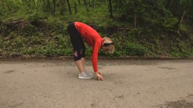 4 k genç sarışın bir kadın Kulaklığımda ormanda çalıştırmadan önce ısınma ve germe gerçekleştirir