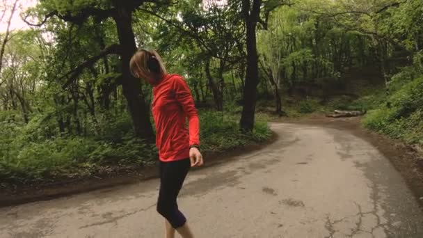 4K jeune femme blonde dans les écouteurs effectue des étirements et le réchauffement avant de courir dans la forêt — Video