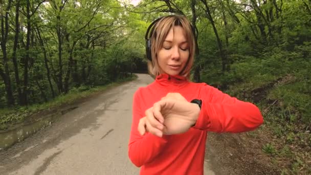 Mujer atractiva joven en auriculares cambiando la configuración de un reloj inteligente en frente de o suela trotando en la carretera en un bosque verde — Vídeos de Stock