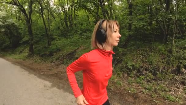 Jeune femme séduisante dans les écouteurs changer les paramètres sur une montre intelligente en face de ou semelle jogging sur la route dans une forêt verte 