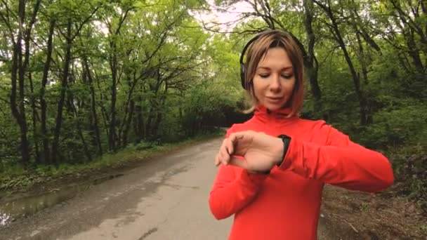 Giovane donna attraente in cuffia cambiare le impostazioni su un orologio intelligente di fronte o suola jogging sulla strada in una foresta verde — Video Stock