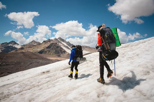 Twee toeristen, een man en een vrouw met rugzakken en stijgijzers op hun voeten lopen langs de gletsjer tegen de achtergrond van de bergen van de lucht en de wolken. Achteraanzicht — Stockfoto