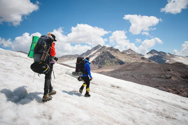 Twee toeristen, een man en een vrouw met rugzakken en stijgijzers op hun voeten lopen langs de gletsjer tegen de achtergrond van de bergen van de lucht en de wolken. Achteraanzicht — Stockfoto