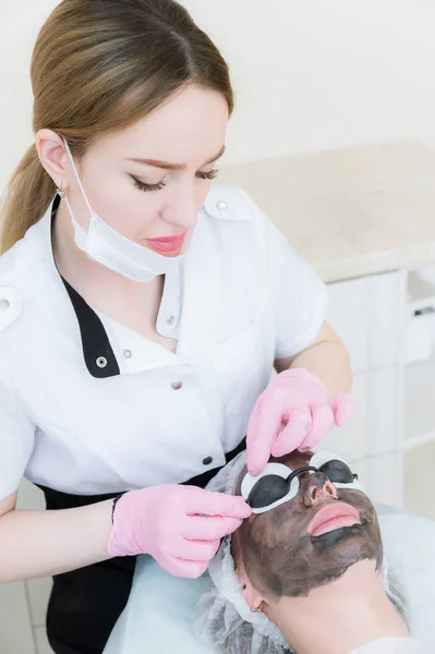 The cosmetologist in pink gloves with a brush applies a carbon mask for peeling on the face of a young girl in a cosmetology room. The concept of cosmetology services and self-care. The concept of