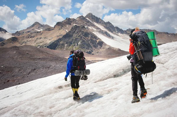 Deux touristes, un homme et une femme avec des sacs à dos et des crampons sur leurs pieds marchent le long du glacier sur le fond des montagnes du ciel et des nuages. Vue arrière — Photo