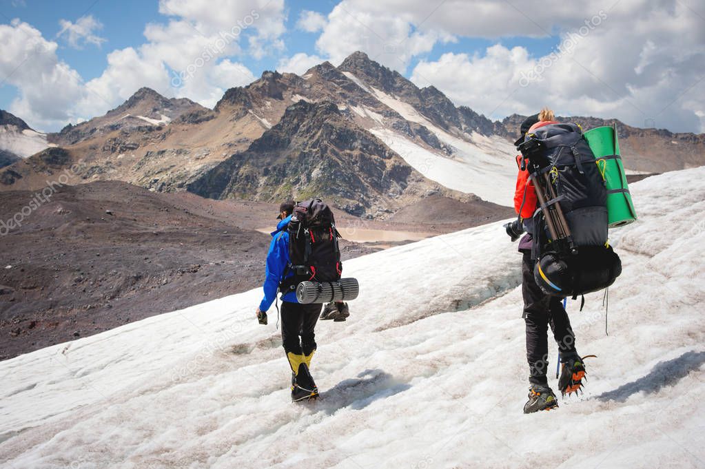 Two tourists, a man and a woman with backpacks and crampons on their feet walk along the glacier against the background of the mountains of the sky and clouds. Back view