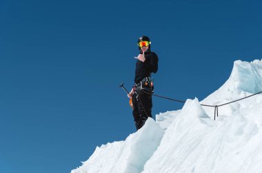 Bir dağcı bir buzulun üzerinde elinde bir kar küreği ile sırtında ve Shaks jest mavi gökyüzüne karşı gösterir