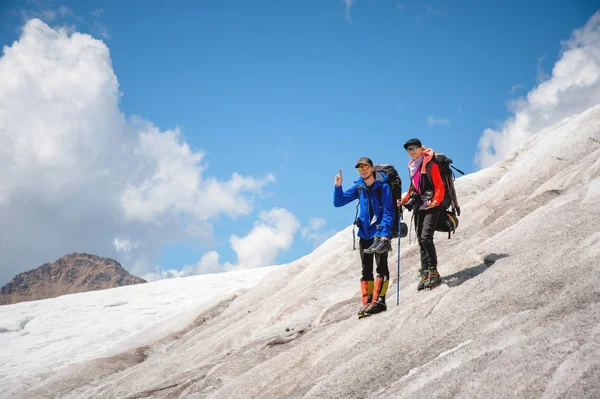 Twee toeristen, een man en een vrouw met rugzakken en katten op hun voeten, staan op het ijs tegen de achtergrond van de bergen van de lucht en de wolken. De man vertoont gebaar kwam het idee — Stockfoto