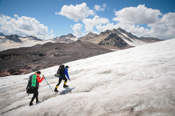 Twee toeristen, een man en een vrouw met rugzakken en stijgijzers op hun voeten lopen langs de gletsjer tegen de achtergrond van de bergen van de lucht en de wolken. Achteraanzicht — Stockfoto