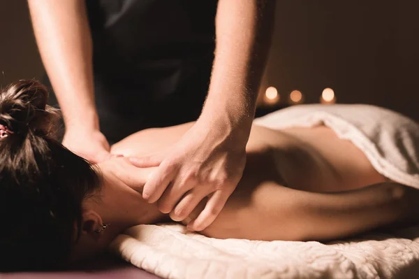 Las manos de hombre hacen un masaje terapéutico en el cuello para una chica acostada en un sofá de masaje en un spa de masaje con iluminación oscura. Primer plano. Llave oscura —  Fotos de Stock