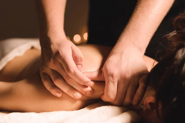 Mens hands make a therapeutic neck massage for a girl lying on a massage couch in a massage spa with dark lighting. Close-up. Dark Key — Stock Photo, Image