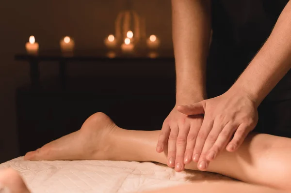 Крупный план мужских рук, делающих телячий массаж женских ног в темной комнате со свечами на заднем плане. Косметология и спа-процедуры — стоковое фото