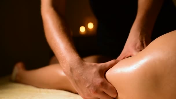 Voor een vrouw doet een masseur in een donkere kamer een hip massage met olie. Spa procedures massage vrouwen — Stockvideo
