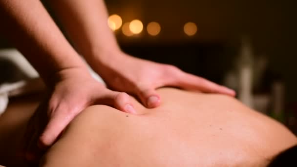 Крупним планом спа-масаж жінок на плечах і спині. Чоловічі руки роблять масаж жінці в темній кімнаті зі свічками на фоні — стокове відео