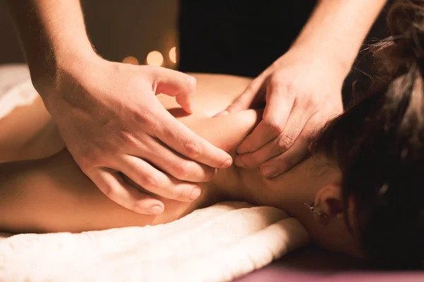 Les mains des hommes font un massage thérapeutique du cou pour une fille allongée sur un canapé de massage dans un spa de massage avec un éclairage sombre. Gros plan. Clé sombre — Photo