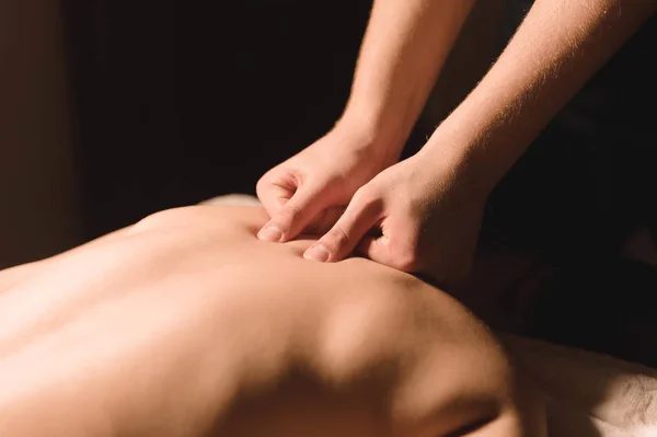 Mens hands make a therapeutic neck massage for a girl lying on a massage couch in a massage spa with dark lighting. Close-up. Dark Key — Stock Photo, Image