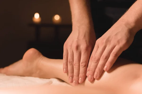 Close-up de mãos masculinas fazendo massagem na panturrilha de pernas femininas em um quarto escuro com velas no fundo. Cosmetologia e tratamentos de spa — Fotografia de Stock