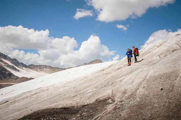 Twee toeristen, een man en een vrouw met rugzakken en katten op hun voeten, staan op het ijs in de achtergrond van de bergen van de lucht en de wolken. communicatie in de bergen — Stockfoto