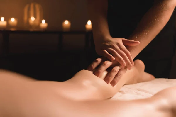 Close-up van mannelijke handen doen kalf massage van vrouwelijke benen in een donkere kamer met kaarsen op de achtergrond. Cosmetologie en wellness-behandelingen — Stockfoto